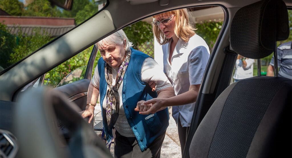 Image of a caretaker helping an elderly woman into the passenger side of a vehicle